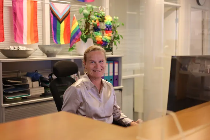 En ansatt hilser velkommen i resepsjonen. Bak henne henger ulike pride-flagg.